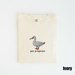 May include: Ivory-colored t-shirt with a crew neck. The shirt features an embroidered cartoon goose holding a cigarette, with the text "got pregnant" below the goose. The shirt is folded neatly on a white surface.
