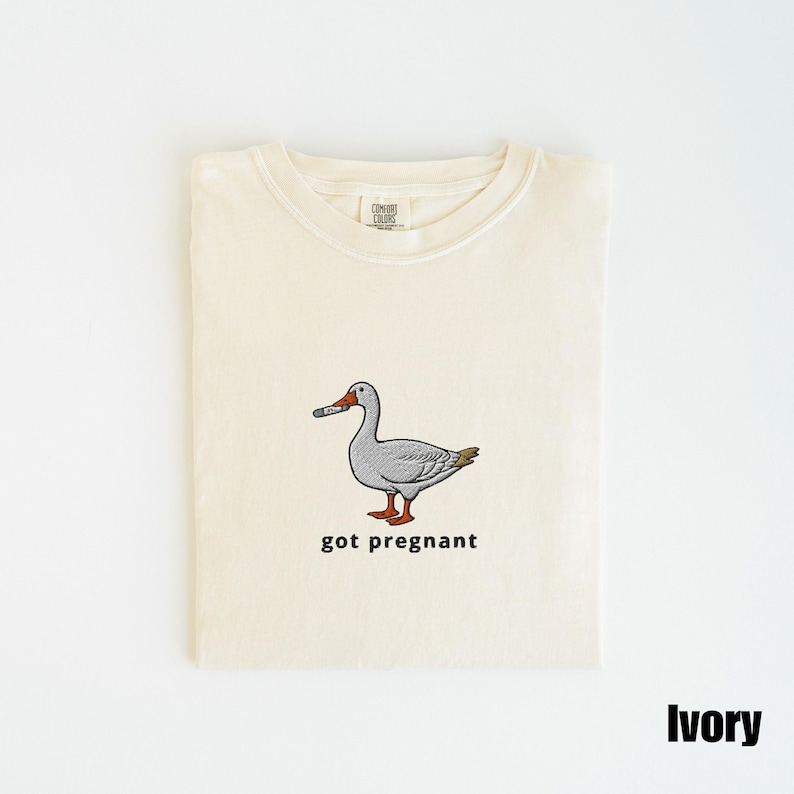 May include: Ivory-colored t-shirt with a crew neck. The shirt features an embroidered cartoon goose holding a cigarette, with the text "got pregnant" below the goose. The shirt is folded neatly on a white surface.