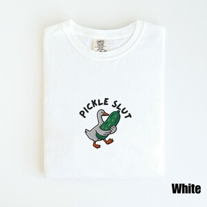 May include: White t-shirt with a crew neck. The shirt features an embroidered design of a gray duck holding a green pickle, with the words "PICKLE SLUT" above the duck. The word "White" is at the bottom right.