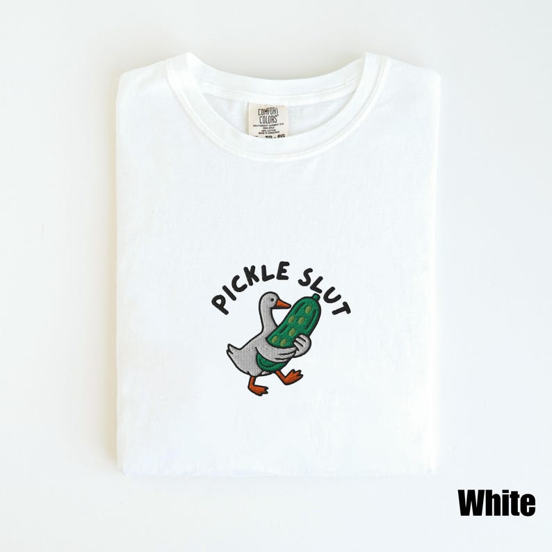 May include: White t-shirt with a crew neck. The shirt features an embroidered design of a gray duck holding a green pickle, with the words "PICKLE SLUT" above the duck. The word "White" is at the bottom right.