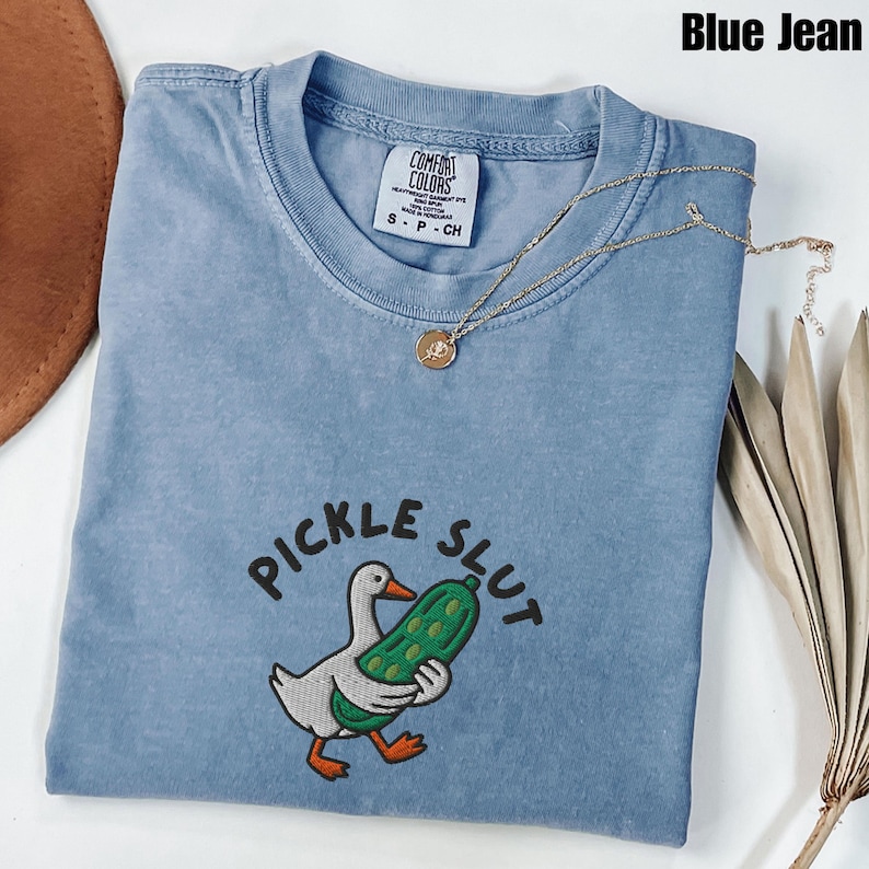 May include: A blue jean Comfort Colors t-shirt with the embroidered phrase "PICKLE SLUT" and a cartoon duck holding a pickle. A gold necklace with a pendant rests on the shirt. The shirt is a size small.