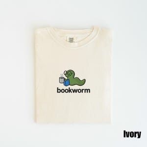 May include: Ivory-colored t-shirt with a crew neck. The shirt features an embroidered design of a green worm wearing glasses, holding a mug and a book, with the word "bookworm" below. The shirt is folded neatly.