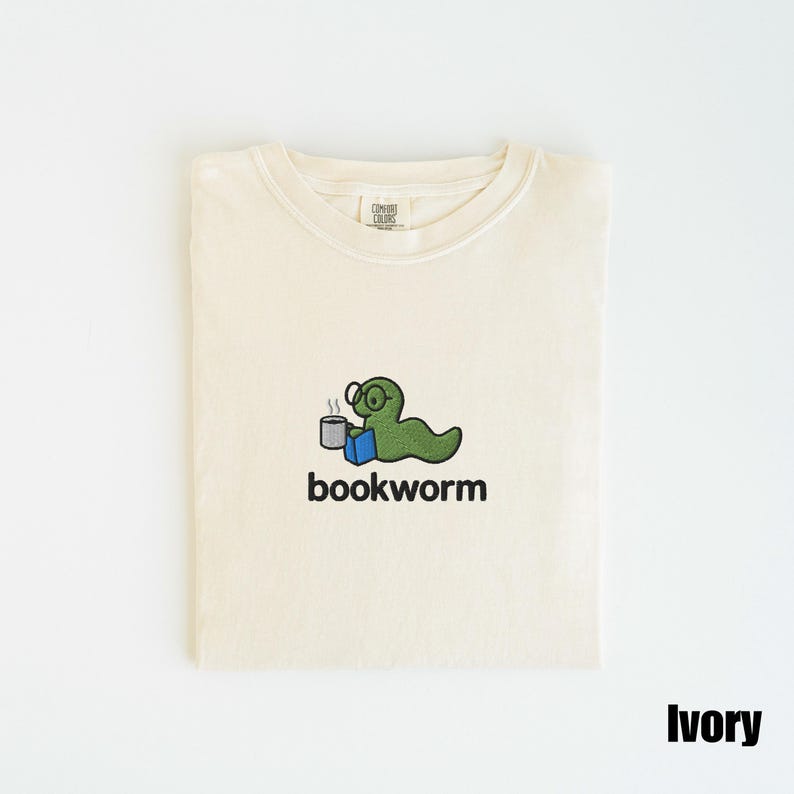 May include: Ivory-colored t-shirt with a crew neck. The shirt features an embroidered design of a green worm wearing glasses, holding a mug and a book, with the word "bookworm" below. The shirt is folded neatly.