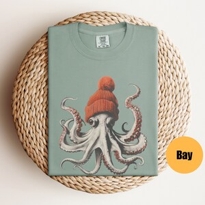 May include: A sage green t-shirt featuring a detailed illustration of an octopus wearing an orange knit cap with a pom-pom. The octopus has large eyes and red-tipped tentacles. The shirt is folded on a woven mat. The tag reads "COMFORT COLORS".