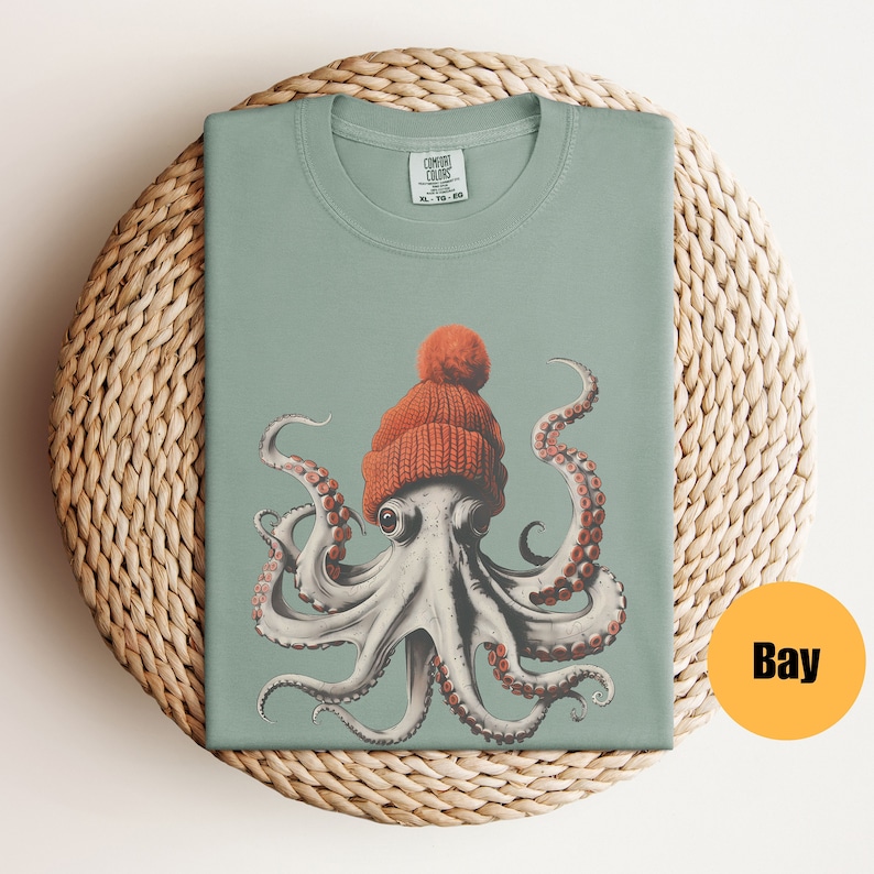 May include: A sage green t-shirt featuring a detailed illustration of an octopus wearing an orange knit cap with a pom-pom. The octopus has large eyes and red-tipped tentacles. The shirt is folded on a woven mat. The tag reads "COMFORT COLORS".