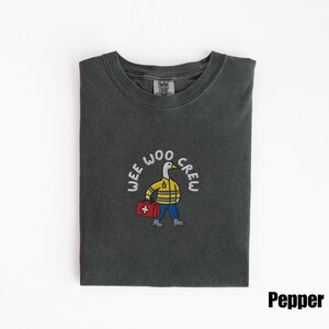 May include: Pepper-colored t-shirt with a crew neck. The shirt features an embroidered design of a goose wearing a yellow jacket and carrying a red first aid kit, with the words "WEE WOO CREW" in a circular pattern.