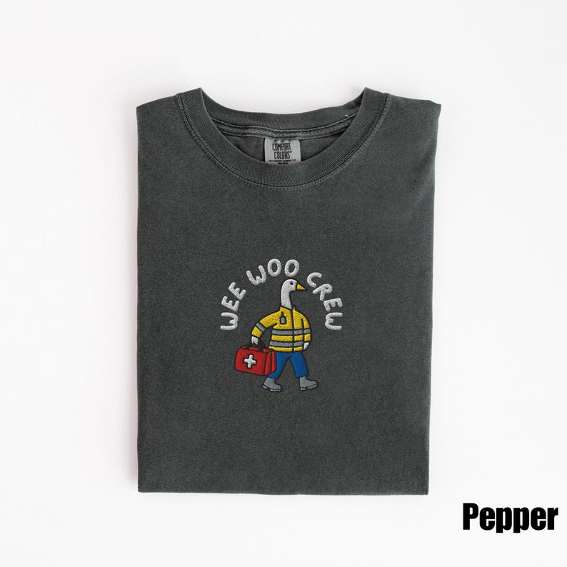 May include: Pepper-colored t-shirt with a crew neck. The shirt features an embroidered design of a goose wearing a yellow jacket and carrying a red first aid kit, with the words "WEE WOO CREW" in a circular pattern.
