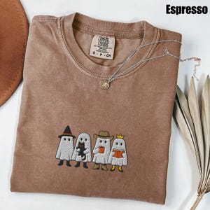 May include: Espresso-colored t-shirt with an embroidered design of four cartoon ghosts, each with a unique accessory. The shirt has a crew neck and a Comfort Colors tag. A silver necklace with a pendant rests on the shirt.