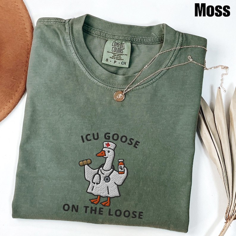 May include: A moss green t-shirt with the text "ICU GOOSE ON THE LOOSE" and an embroidered goose wearing a nurse's uniform. A gold necklace with a pendant is draped across the shirt. The shirt has a Comfort Colors tag.