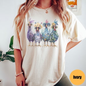 Peut inclure: Un t-shirt en ivoire clair avec un motif de quatre poulets portant des lunettes de soleil et des bandeaux floraux. Les poulets sont align&eacute;s, face &agrave; l'avant. Le t-shirt est de coupe d&eacute;contract&eacute;e et a un col rond.