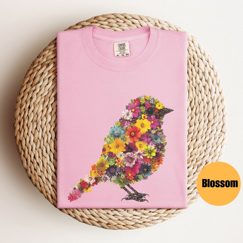 May include: Pink t-shirt with a floral bird design. The bird is made up of colorful flowers and has a black beak and eyes. The shirt has a tag that says "Comfort Colors".
