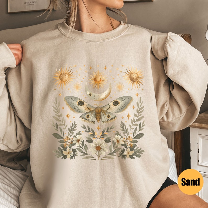 Pode incluir: Uma camisa de moletom bege com uma estampa gr&aacute;fica de uma mariposa, lua e s&oacute;is cercados por flores e estrelas. O texto "Sand" &eacute; vis&iacute;vel no canto inferior direito da imagem.