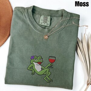 Pu&ograve; includere: T-shirt verde muschio con ricamo di una rana che tiene un calice di vino. La rana ha un fiore viola e rossetto rosso. Una collana d'oro con un ciondolo &egrave; sulla maglietta. La maglietta ha un'etichetta Comfort Colors.