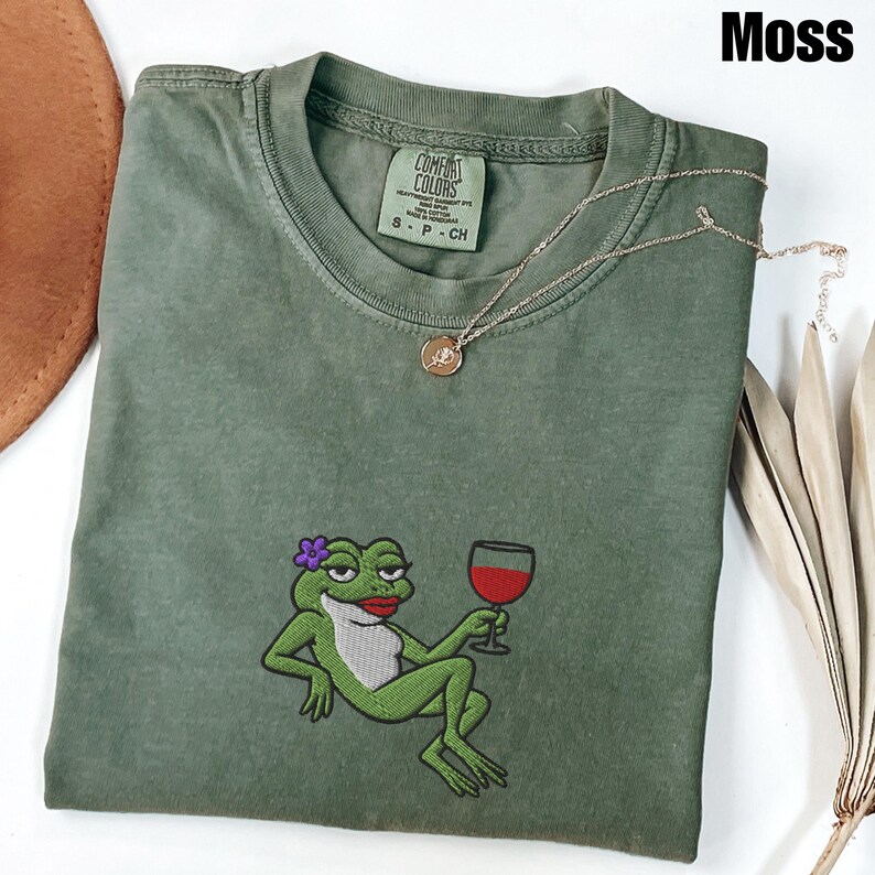 Pu&ograve; includere: T-shirt verde muschio con ricamo di una rana che tiene un calice di vino. La rana ha un fiore viola e rossetto rosso. Una collana d'oro con un ciondolo &egrave; sulla maglietta. La maglietta ha un'etichetta Comfort Colors.
