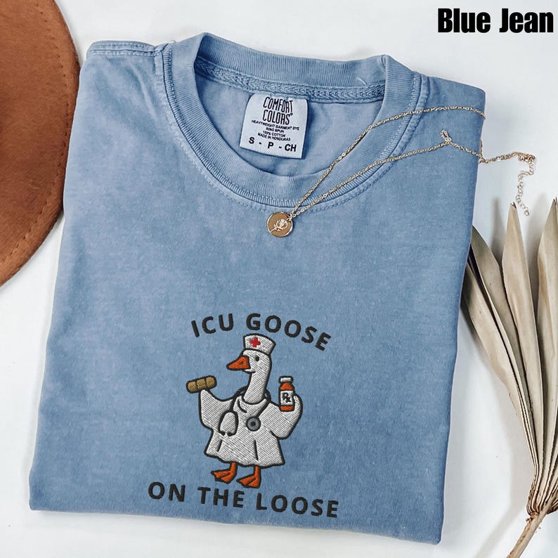 May include: A blue jean Comfort Colors t-shirt with the text "ICU Goose ON THE LOOSE" and an embroidered cartoon goose wearing a doctor's coat. A gold necklace with a pendant is draped across the shirt.