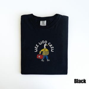 May include: Black t-shirt with the embroidered text "WEE WOO CREW" above a cartoon duck wearing a yellow jacket, blue pants, and carrying a red medical bag. The shirt is folded neatly.