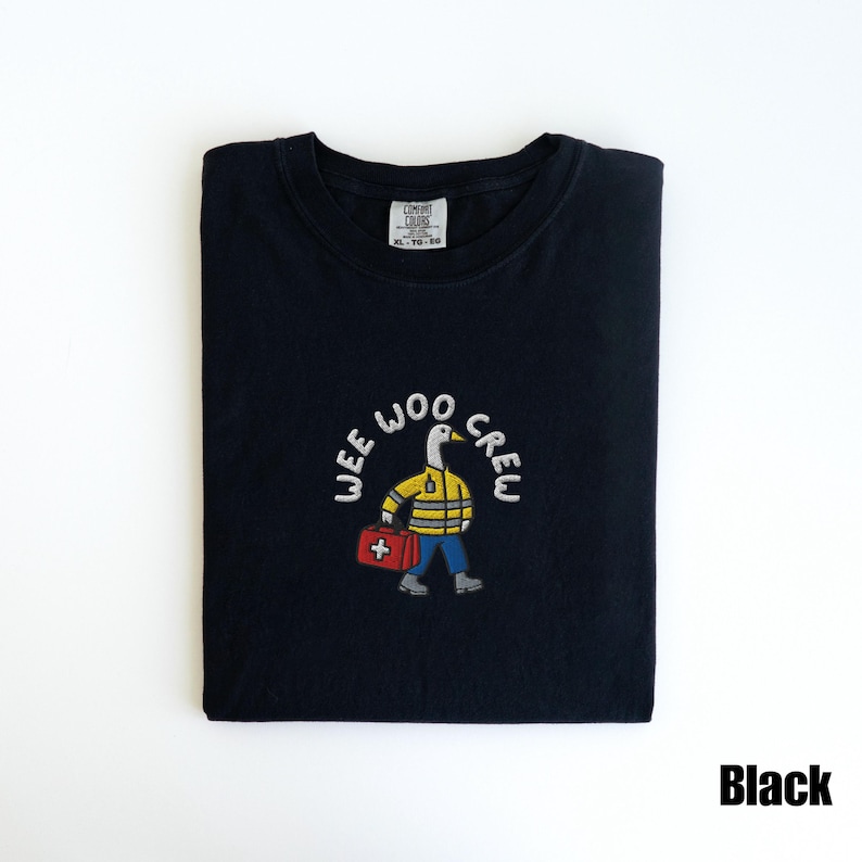 May include: Black t-shirt with the embroidered text "WEE WOO CREW" above a cartoon duck wearing a yellow jacket, blue pants, and carrying a red medical bag. The shirt is folded neatly.