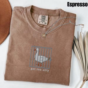May include: A brown t-shirt with a white embroidered goose in a cage and the text "got too silly". The shirt is on a brown surface with a silver necklace and a brown hat.