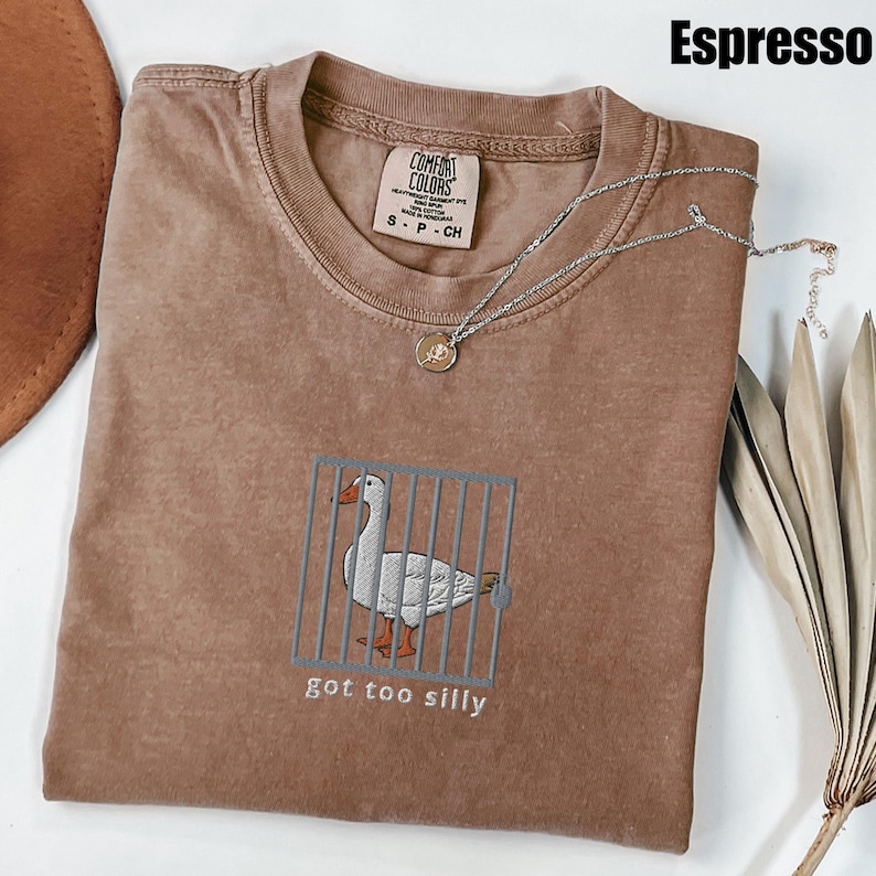 May include: A brown t-shirt with a white embroidered goose in a cage and the text "got too silly". The shirt is on a brown surface with a silver necklace and a brown hat.