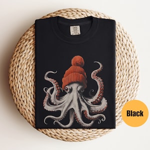 May include: Black t-shirt featuring a detailed illustration of an octopus wearing a knitted orange beanie with a pom-pom. The octopus has gray skin and red suckers on its tentacles. The shirt is folded on a woven, circular surface.