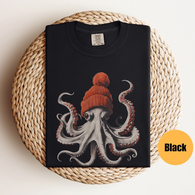 May include: Black t-shirt featuring a detailed illustration of an octopus wearing a knitted orange beanie with a pom-pom. The octopus has gray skin and red suckers on its tentacles. The shirt is folded on a woven, circular surface.