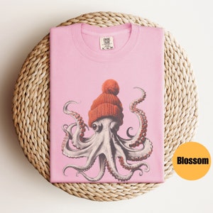 May include: A light pink t-shirt featuring a detailed illustration of an octopus wearing an orange knit cap with a pom-pom. The shirt is folded on a woven, circular placemat. The tag reads "COMFORT COLORS". The word "Blossom" is in a yellow circle.