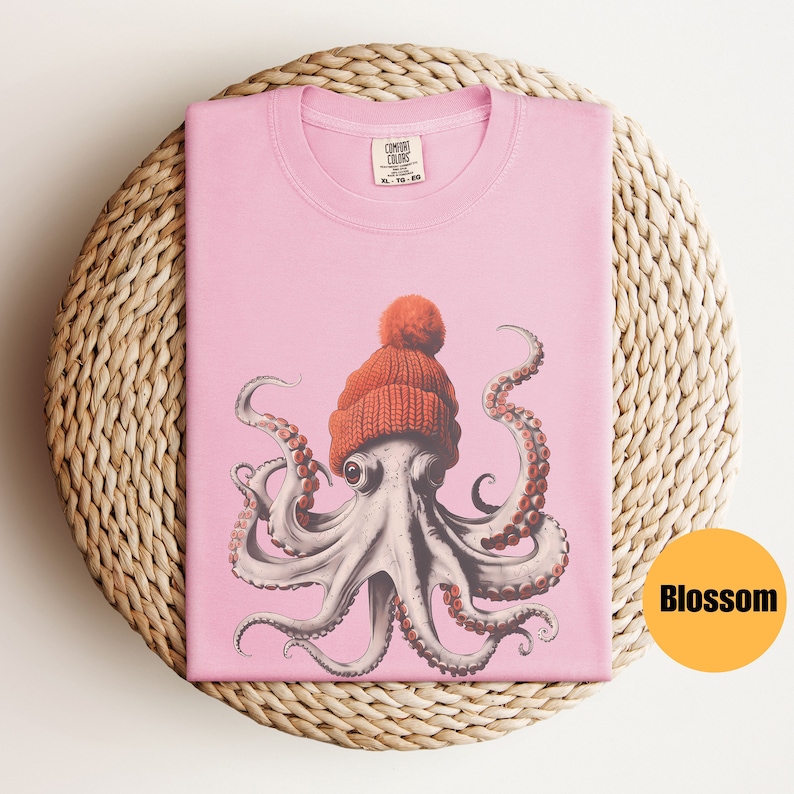 May include: A light pink t-shirt featuring a detailed illustration of an octopus wearing an orange knit cap with a pom-pom. The shirt is folded on a woven, circular placemat. The tag reads "COMFORT COLORS". The word "Blossom" is in a yellow circle.