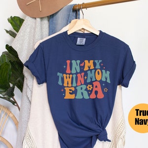 Może przedstawiać: Koszulka w kolorze granatowym z napisem "In My Twin Mom Era" w retro czcionce tęczowej. Koszulka wisi na drewnianym wieszaku.