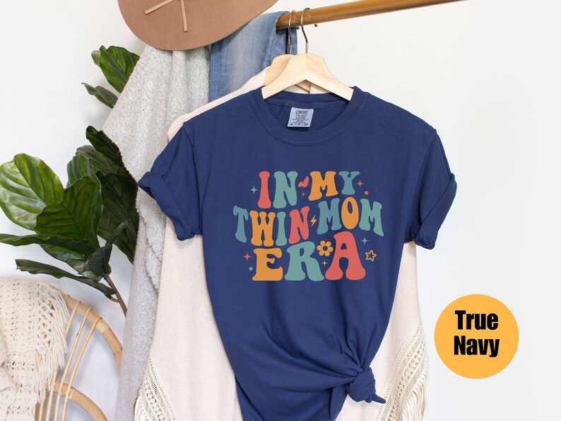 Może przedstawiać: Koszulka w kolorze granatowym z napisem "In My Twin Mom Era" w retro czcionce tęczowej. Koszulka wisi na drewnianym wieszaku.