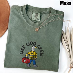 May include: A moss green Comfort Colors t-shirt with the words "WEE WOO CREW" embroidered on the front. The graphic features a cartoon duck wearing a yellow jacket, blue pants, and carrying a red medical bag. A gold necklace with a pendant is draped on the shirt.