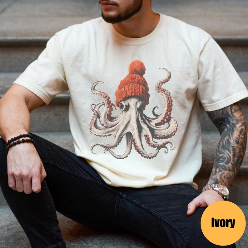 May include: Ivory-colored t-shirt featuring a detailed illustration of an octopus wearing an orange knit beanie. The octopus has multiple tentacles and is set against a neutral background. The shirt is a casual style.