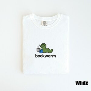 May include: White t-shirt featuring an embroidered design of a green worm wearing glasses, holding a mug and a blue book. The word "bookworm" is below the graphic. The shirt has a crew neck and the word "White" is printed at the bottom.