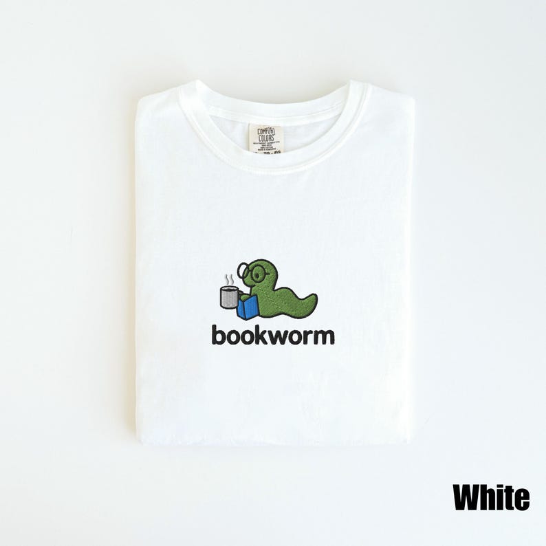 May include: White t-shirt featuring an embroidered design of a green worm wearing glasses, holding a mug and a blue book. The word "bookworm" is below the graphic. The shirt has a crew neck and the word "White" is printed at the bottom.