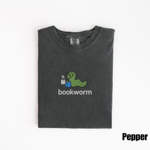May include: Pepper-colored t-shirt with a crew neck. The shirt features a green cartoon bookworm wearing glasses and holding a cup of coffee, with the word "bookworm" below. The shirt is folded neatly.