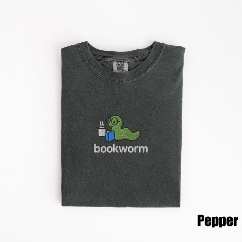 May include: Pepper-colored t-shirt with a crew neck. The shirt features a green cartoon bookworm wearing glasses and holding a cup of coffee, with the word "bookworm" below. The shirt is folded neatly.