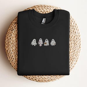 May include: A black crewneck sweatshirt featuring an embroidered design of four white ghosts in the centre. Each ghost has a distinct pattern, including floral, polka dot, and pumpkin motifs, rendered in white, pink, and orange thread.