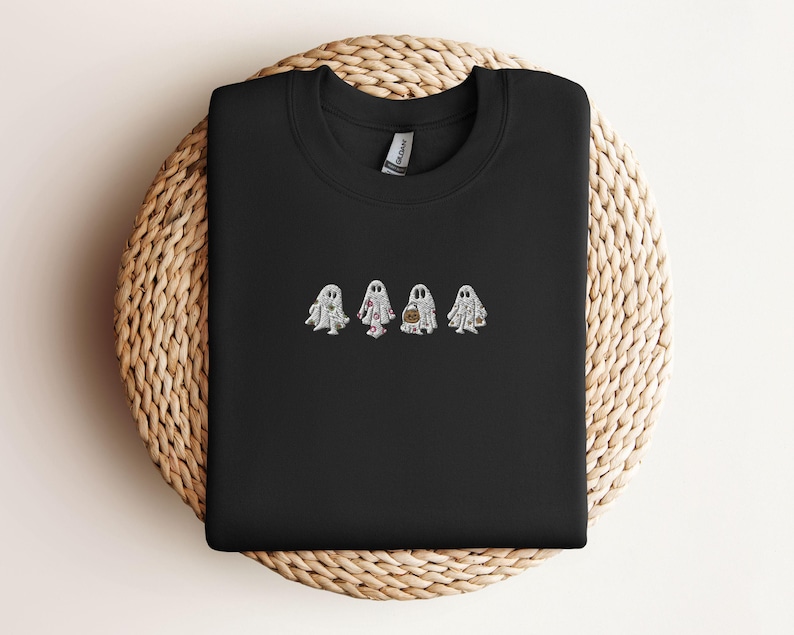 May include: A black crewneck sweatshirt featuring an embroidered design of four white ghosts in the centre. Each ghost has a distinct pattern, including floral, polka dot, and pumpkin motifs, rendered in white, pink, and orange thread.