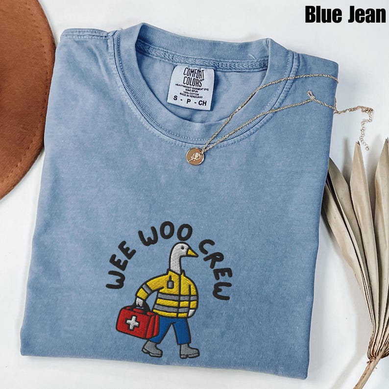 May include: A blue jean Comfort Colors t-shirt with the text "Wee Woo Crew" and a cartoon duck wearing a yellow jacket and carrying a red medical bag. A gold necklace with a pendant is draped across the shirt.