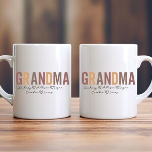 以下が含まれることがあります： ブラウン、オレンジ、ピンクの文字で「Grandma」と書かれた白いセラミック製のマグカップ2つ。マグカップには、「Zachary、Allison、Logan、Camden、Lucas」という名前が「Grandma」の下に書かれており、各名前の間にハートマークがあります。