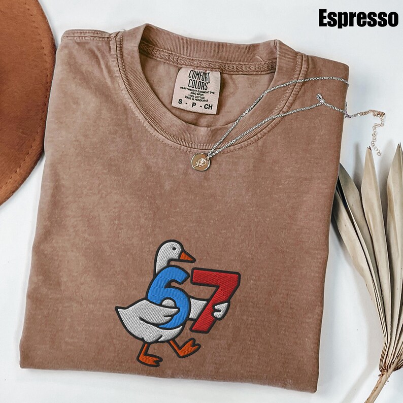 Peut inclure: T-shirt couleur expresso avec un dessin de canard tenant les chiffres 6 et 7. Le t-shirt a un col rond et une &eacute;tiquette Comfort Colors. Un collier en argent avec un pendentif est drap&eacute; sur le t-shirt.