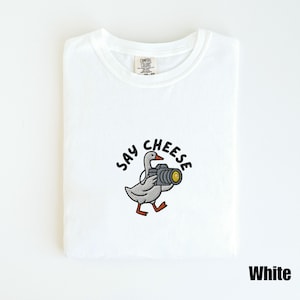 May include: White t-shirt with a crew neck. The shirt features an embroidered design of a goose holding a camera, with the words "Say Cheese" above the goose. The word "White" is printed in black at the bottom right.