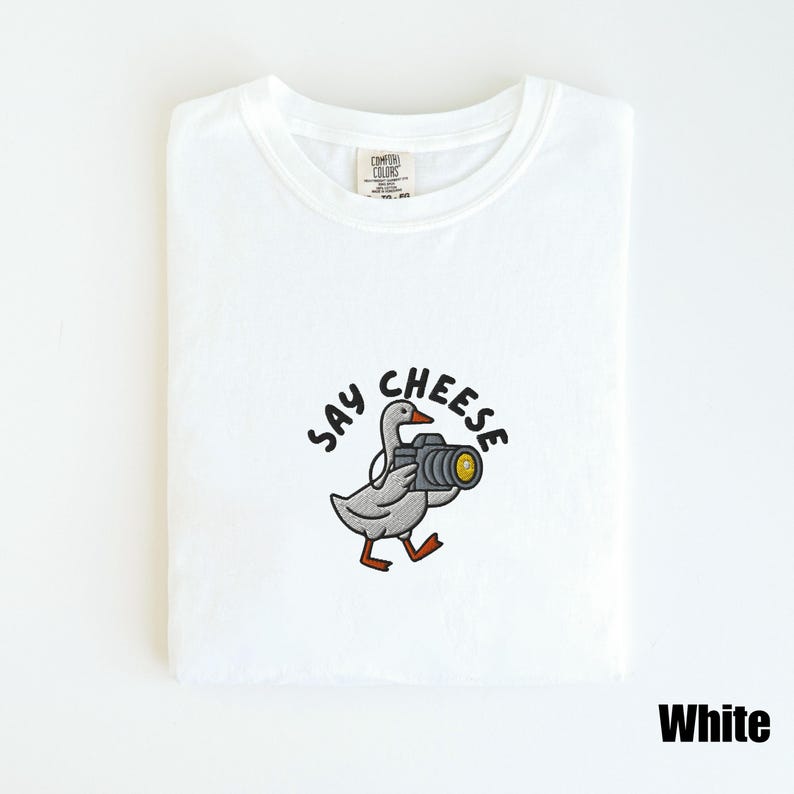 May include: White t-shirt with a crew neck. The shirt features an embroidered design of a goose holding a camera, with the words "Say Cheese" above the goose. The word "White" is printed in black at the bottom right.