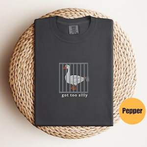 May include: A dark gray t-shirt with a white goose in a cage and the text "got too silly".