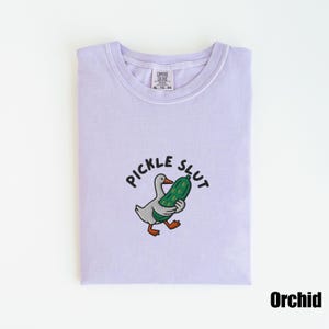 May include: A light purple t-shirt with the text "PICKLE SLUT" above an embroidered cartoon duck holding a pickle. The shirt is labeled "Orchid" and has a Comfort Colors tag.