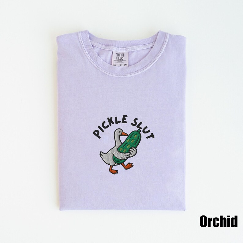 May include: A light purple t-shirt with the text "PICKLE SLUT" above an embroidered cartoon duck holding a pickle. The shirt is labeled "Orchid" and has a Comfort Colors tag.