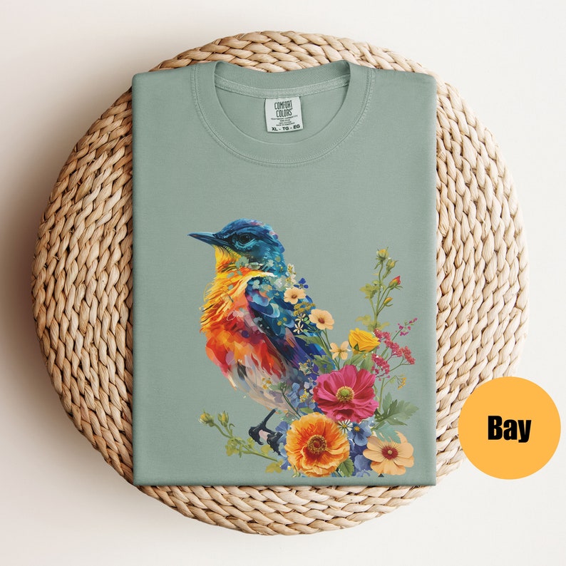 May include: A teal blue, orange, and yellow bird with a long beak sits on a bed of colourful flowers. The bird is surrounded by pink, yellow, and blue flowers. The image is printed on a light green t-shirt.