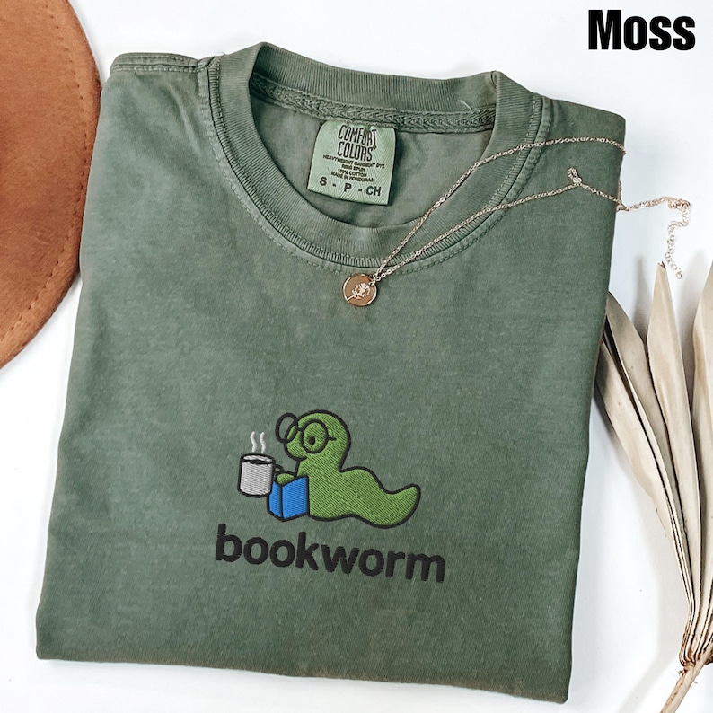 May include: A moss green t-shirt with a graphic of a green worm wearing glasses, holding a coffee cup and a blue book. The word "bookworm" is printed below the graphic. A gold necklace with a pendant is on the shirt.