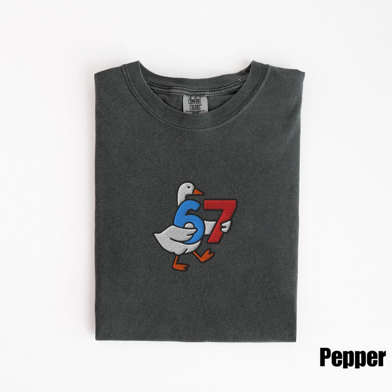 Peut inclure: T-shirt gris fonc&eacute; avec un motif brod&eacute; d'une oie portant les chiffres 6 et 7. L'oie est blanche avec les pieds et le bec orange. Les chiffres sont bleus et rouges. Le mot "Pepper" est dans le coin inf&eacute;rieur droit.
