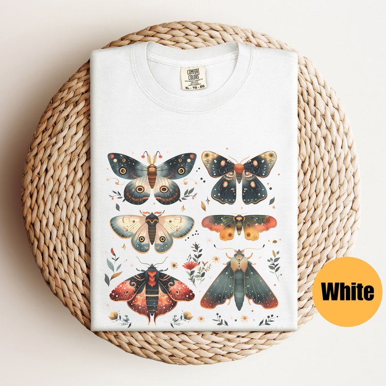 Puede incluir: Camiseta blanca con un gr&aacute;fico de seis polillas coloridas con detalles intrincados y acentos florales. Las polillas tienen un tema celestial con estrellas y lunas en sus alas.