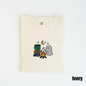 May include: Ivory-colored t-shirt with an embroidered design of a Frankenstein-like character and a ghost roasting marshmallows over a campfire under a crescent moon and stars. The shirt is folded neatly.